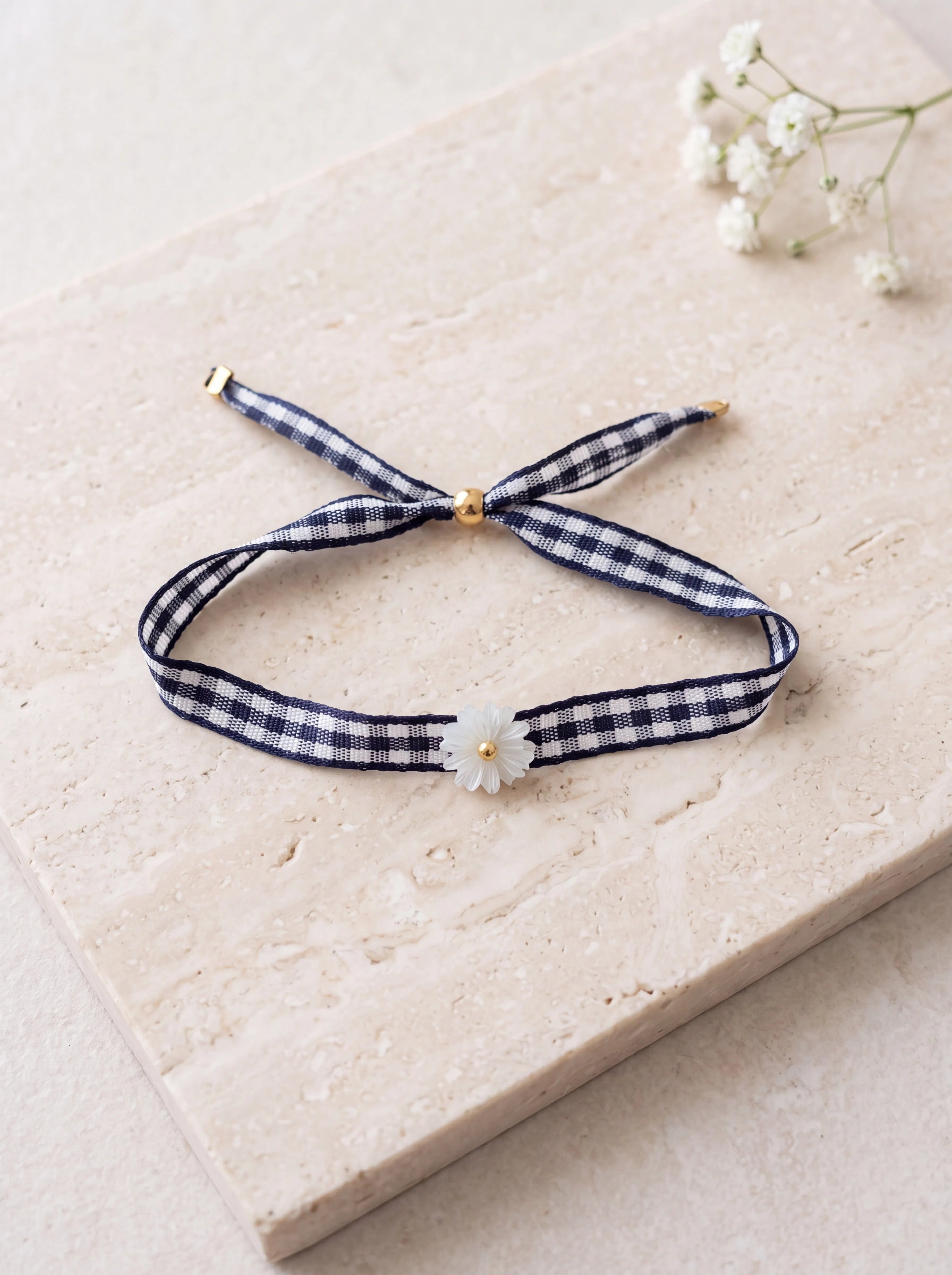 Bracelet LISON en ruban vichy bleu marine avec fleur en nacre et finitions dorées à l'or 24 carats sur support en pierre.