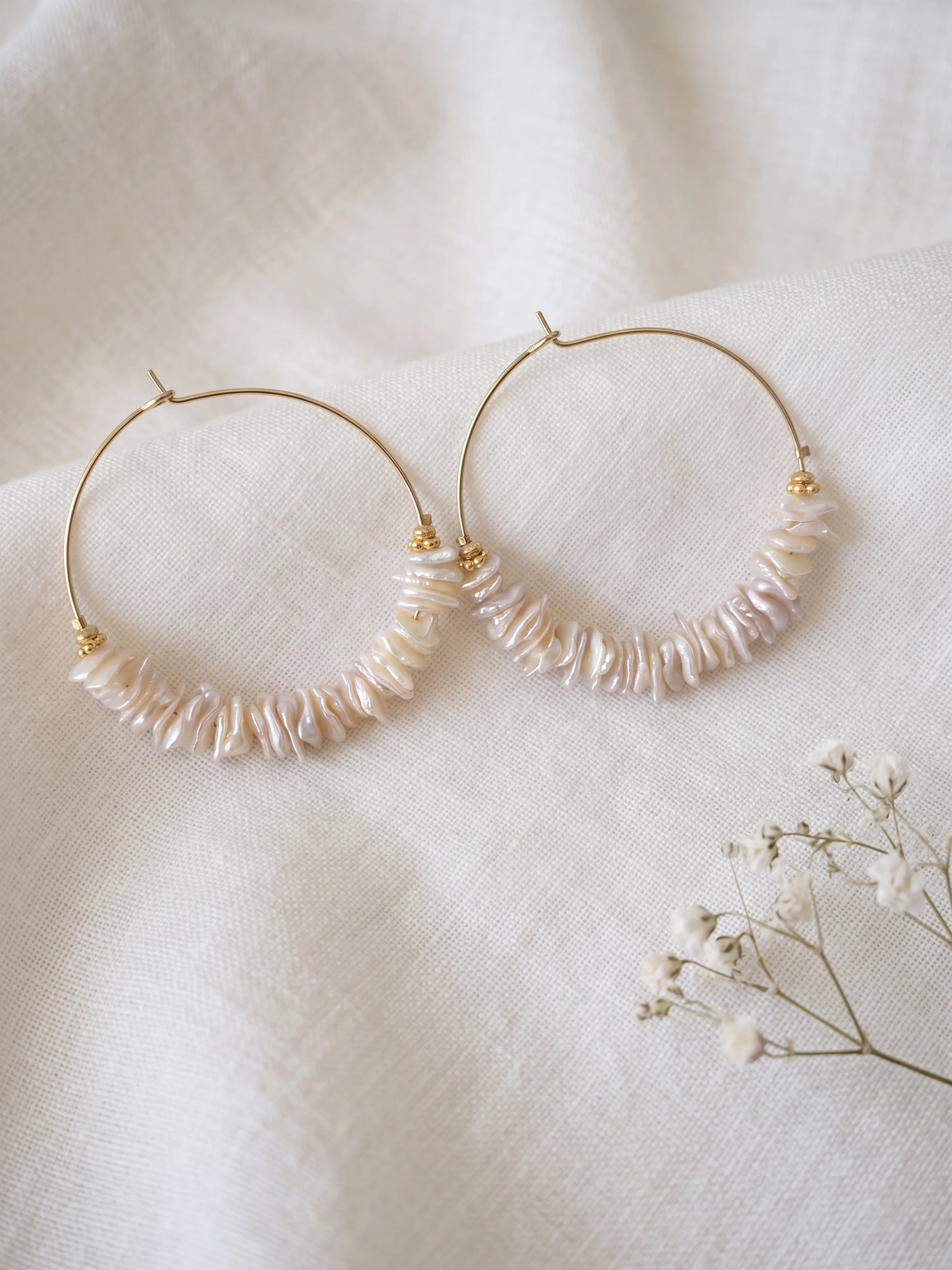 Créoles dorées de 40 mm ornées de perles de nacre irrégulières et de petites perles dorées sur fond de lin blanc.